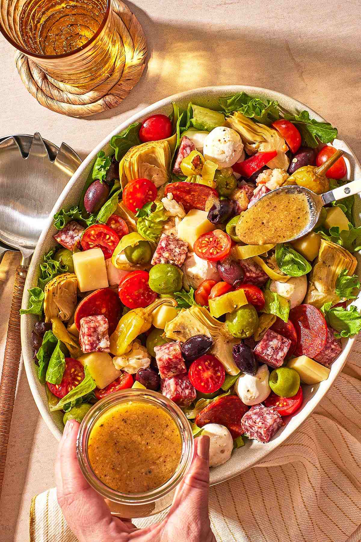 an overhead photo of dijon vinaigrette being spooned from a jar onto the antipasto salad, next to a glass of water and serving utensils.