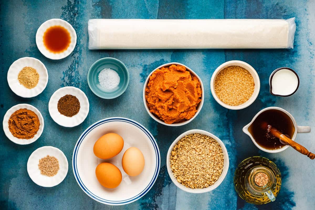 ingredients for baklava pumpkin pie including pumpkin puree, eggs, honey, raw cane sugar, whole milk, vanilla extract, cinnamon, ground ginger, allspice, nutmeg, walnuts, phyllo dough, and olive oil.