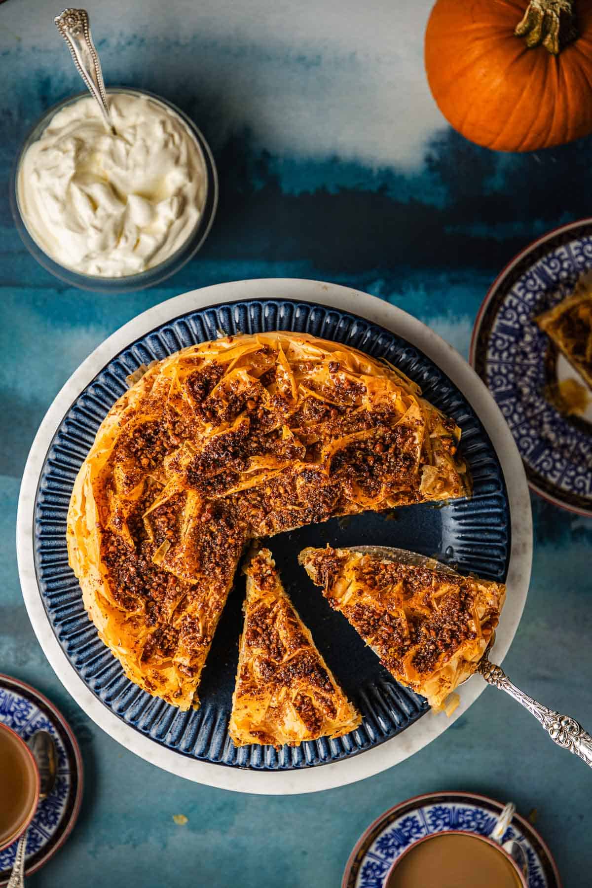 an overhead photo of a baklava pumpkin pie with two slices cut surrounded by cups of coffee, a pumpkin, and a small bowl of whipped cream with a spoon.