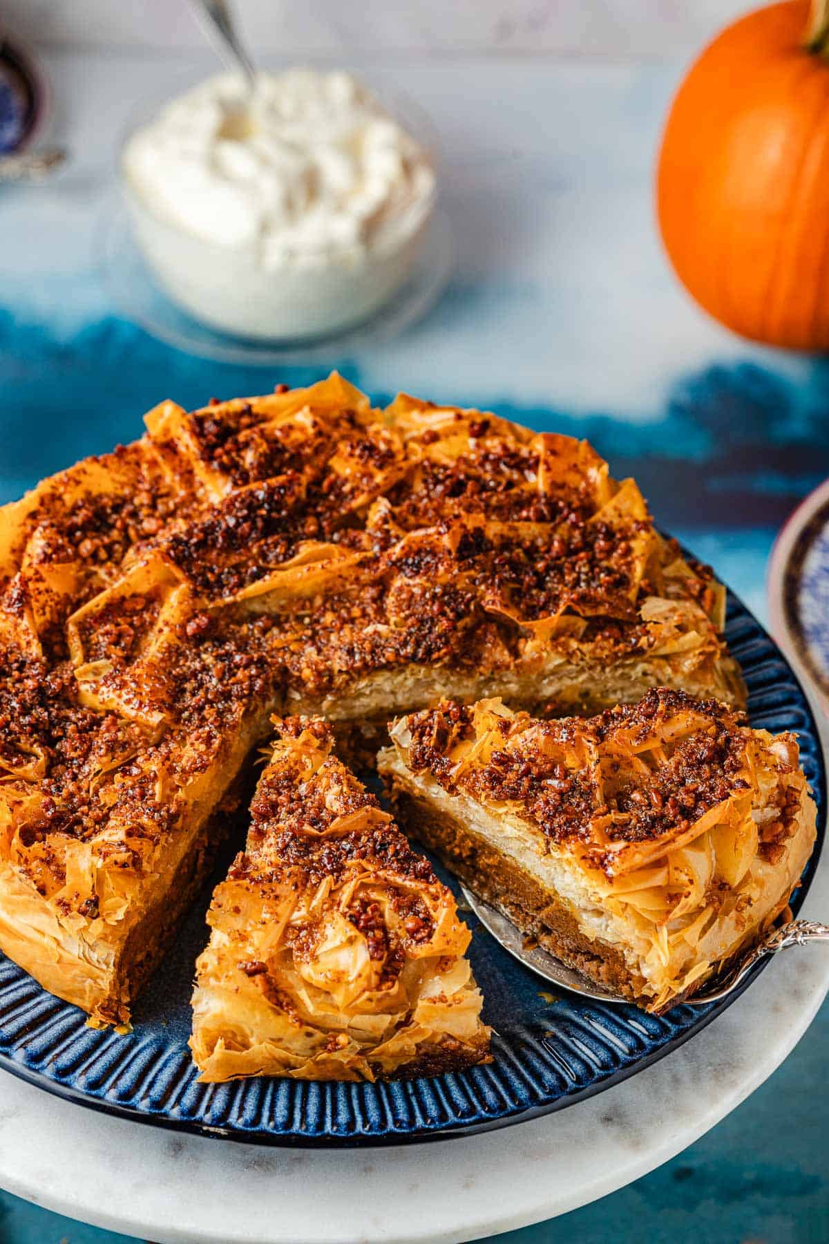 a close up of a baked baklava pumpkin pie with two slices cut into it, one being lifted out with a pie server with a small bowl of whipped cream and a pumpkin in the background.