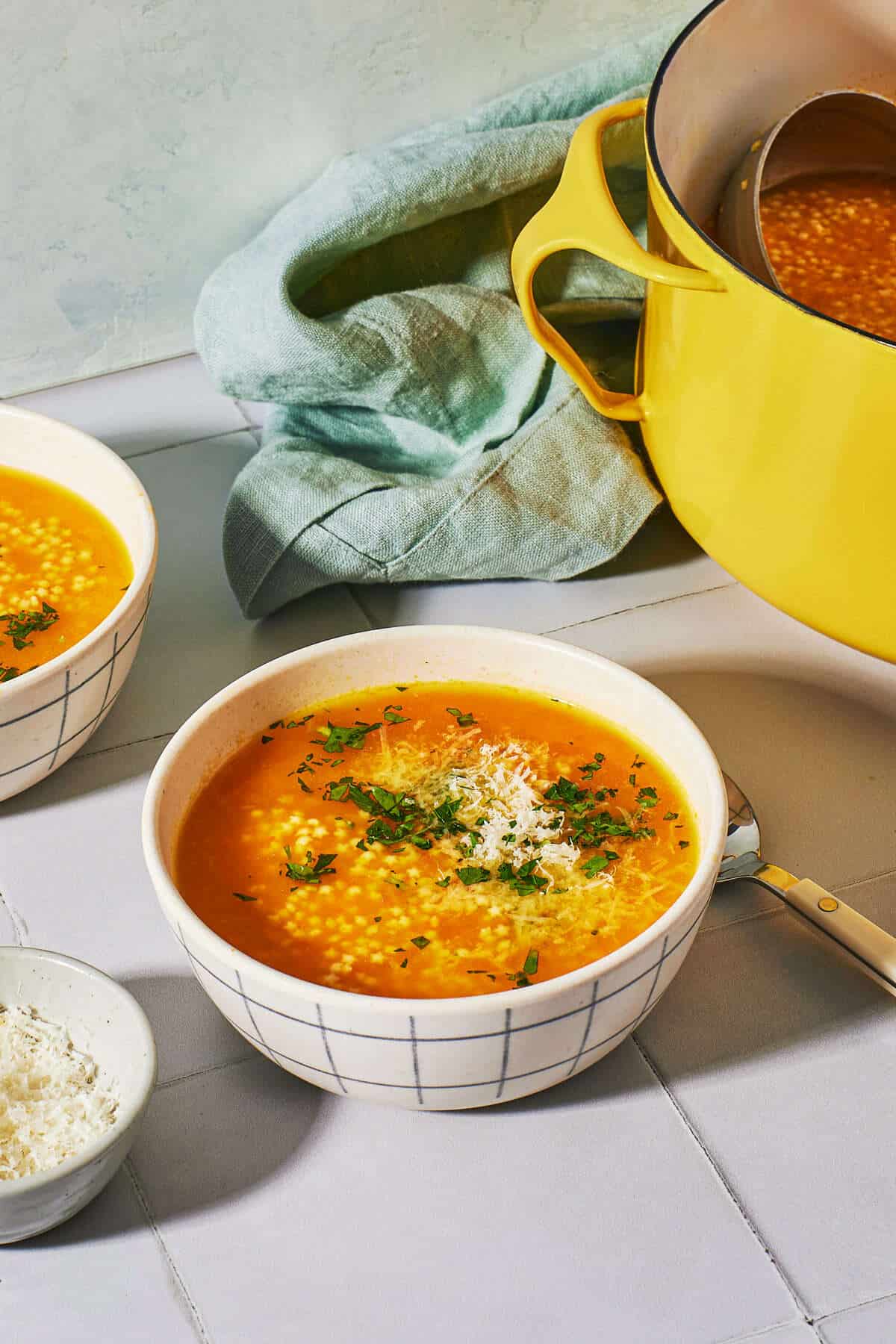 a bowl of pastina soup next to a spoon, another bowl of soup, a pot of soup and a small bowl of grated parmesan cheese.