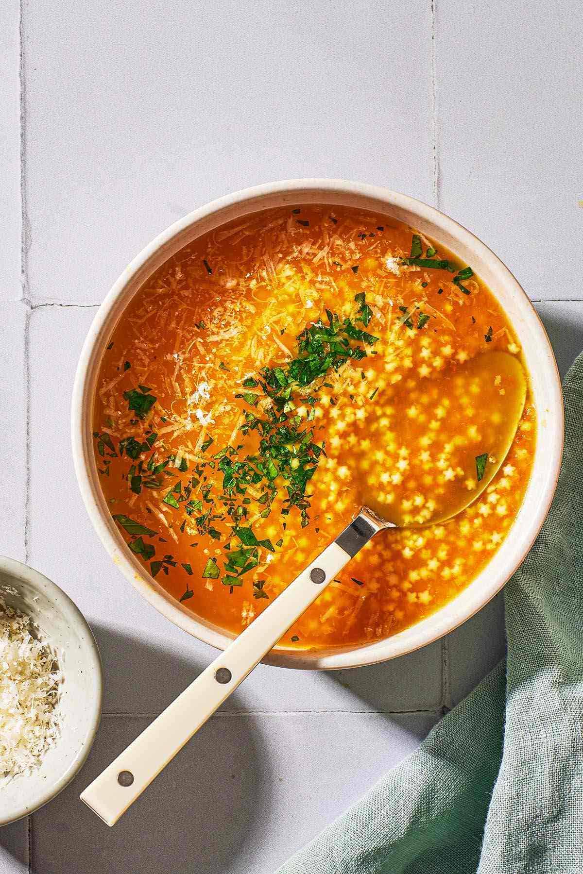 an overhead photo of a serving of pastina soup garnished with parsley and parmesan cheese in a bowl with a spoon.