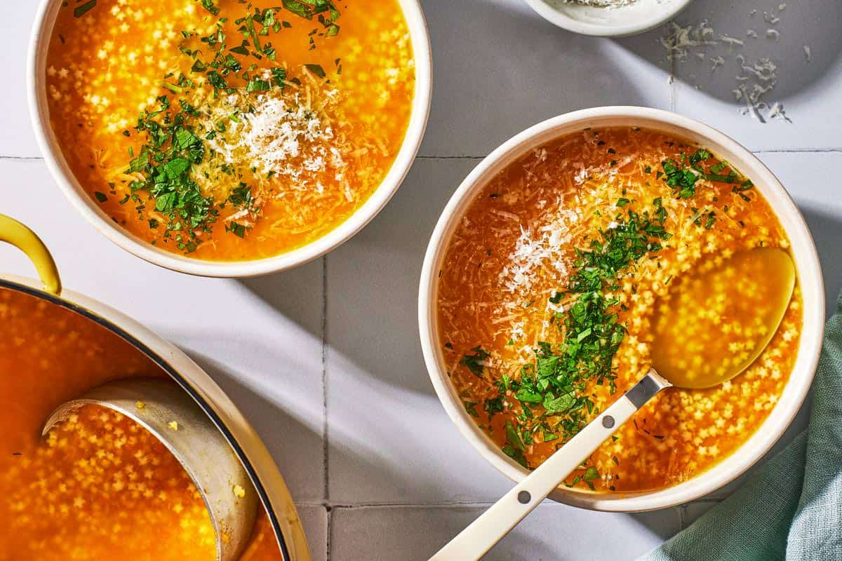 an overhead photo of 2 bowls of pastina soup garnished with parsley and parmesan cheese, one with a spoon, next to a pot of soup with a ladle.