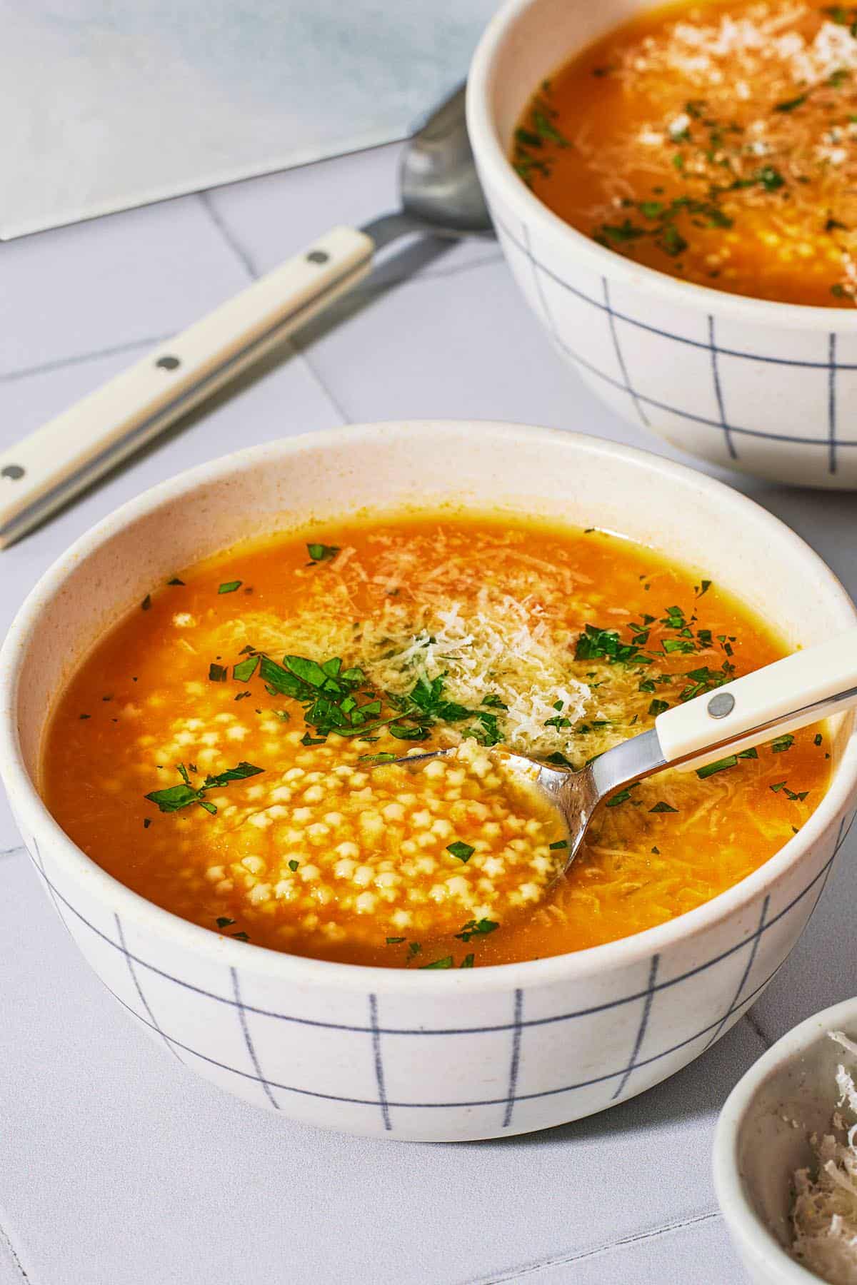 2 bowls of pastina soup garnished with parsley and parmesan cheese, one with a spoon in it, one with a spoon next to it.