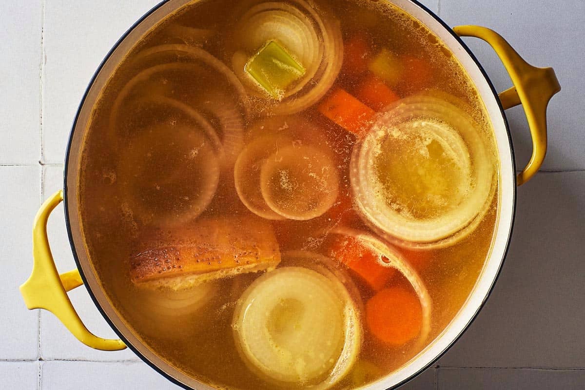 an overhead photo of a pot of simmering broth for pastina soup with the onion, carrots, celery and parmesan rind.