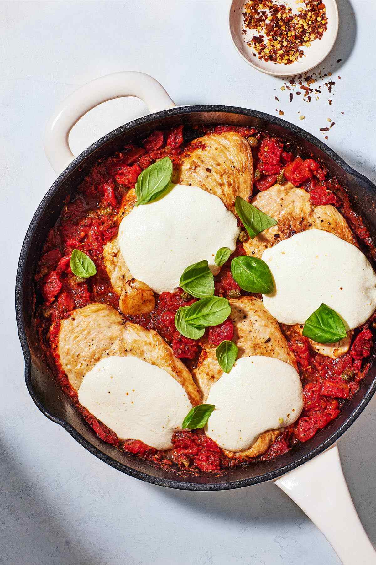 pollo alla pizzaiola chicken in tomato sauce in a skillet next to a small bowl of red pepper flakes.