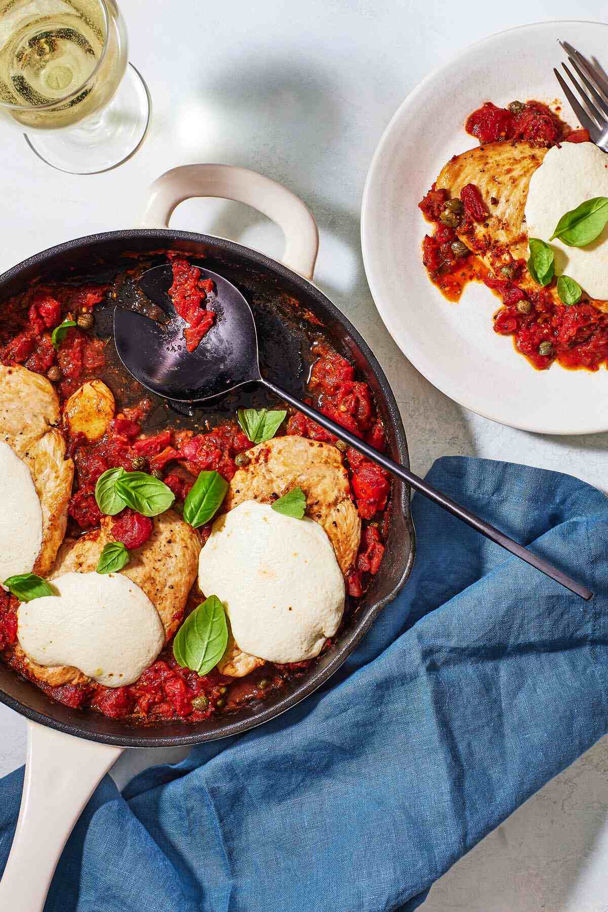 a glass of wine and a skillet of pollo alla pizzaiola chicken in tomato sauce, with a portion removed, sitting on a plate next to it.