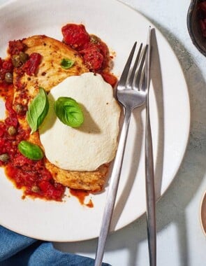 a serving of pollo alla pizzaiola chicken in tomato sauce on a plate with a knife and fork next to a skillet of the chicken and a small bowl of red pepper flakes.