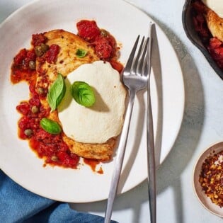 a serving of pollo alla pizzaiola chicken in tomato sauce on a plate with a knife and fork next to a skillet of the chicken and a small bowl of red pepper flakes.