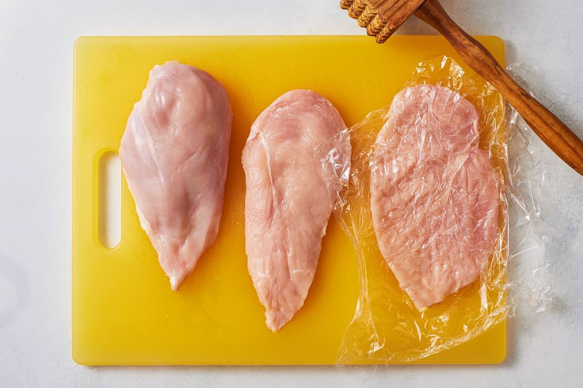3 chicken cutlets on a cutting board, one covered with plastic wrap and flattened with a nearby mallet.