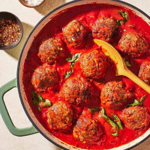 meatballs in sauce in a large pot with a wooden spoon next to small bowls of red pepper flakes and grated parmesan cheese.