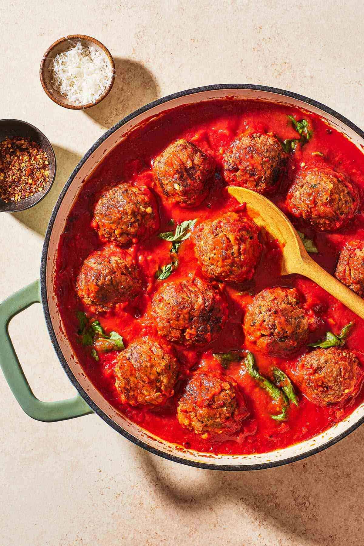 meatballs in sauce in a large pot with a wooden spoon next to small bowls of red pepper flakes and grated parmesan cheese.