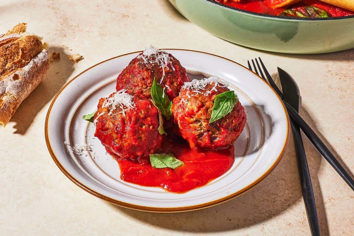 three meatballs and sauce garnished with basil and grated cheese on a plate next to a knife and fork and some crusty bread.