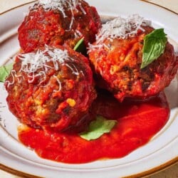a close up of three meatballs and sauce garnished with basil and grated cheese on a plate next to some crusty bread.