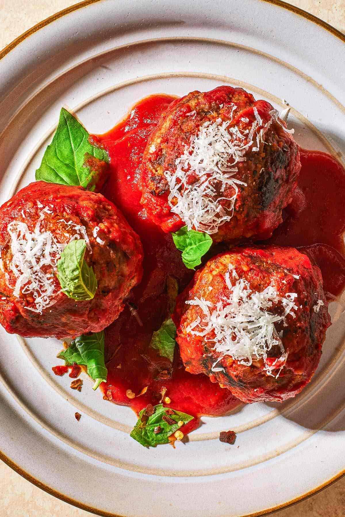 an overhead photo of three meatballs and sauce garnished with basil and grated cheese on a plate.