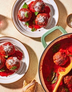 an overhead photo of two plates of meatballs and sauce, a pot of meatballs with a wooden spoon, a knife and fork, 2 drinks and a small bowl of grated cheese.