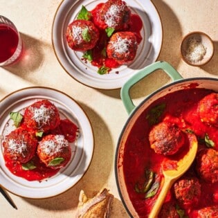 an overhead photo of two plates of meatballs and sauce, a pot of meatballs with a wooden spoon, a knife and fork, 2 drinks and a small bowl of grated cheese.
