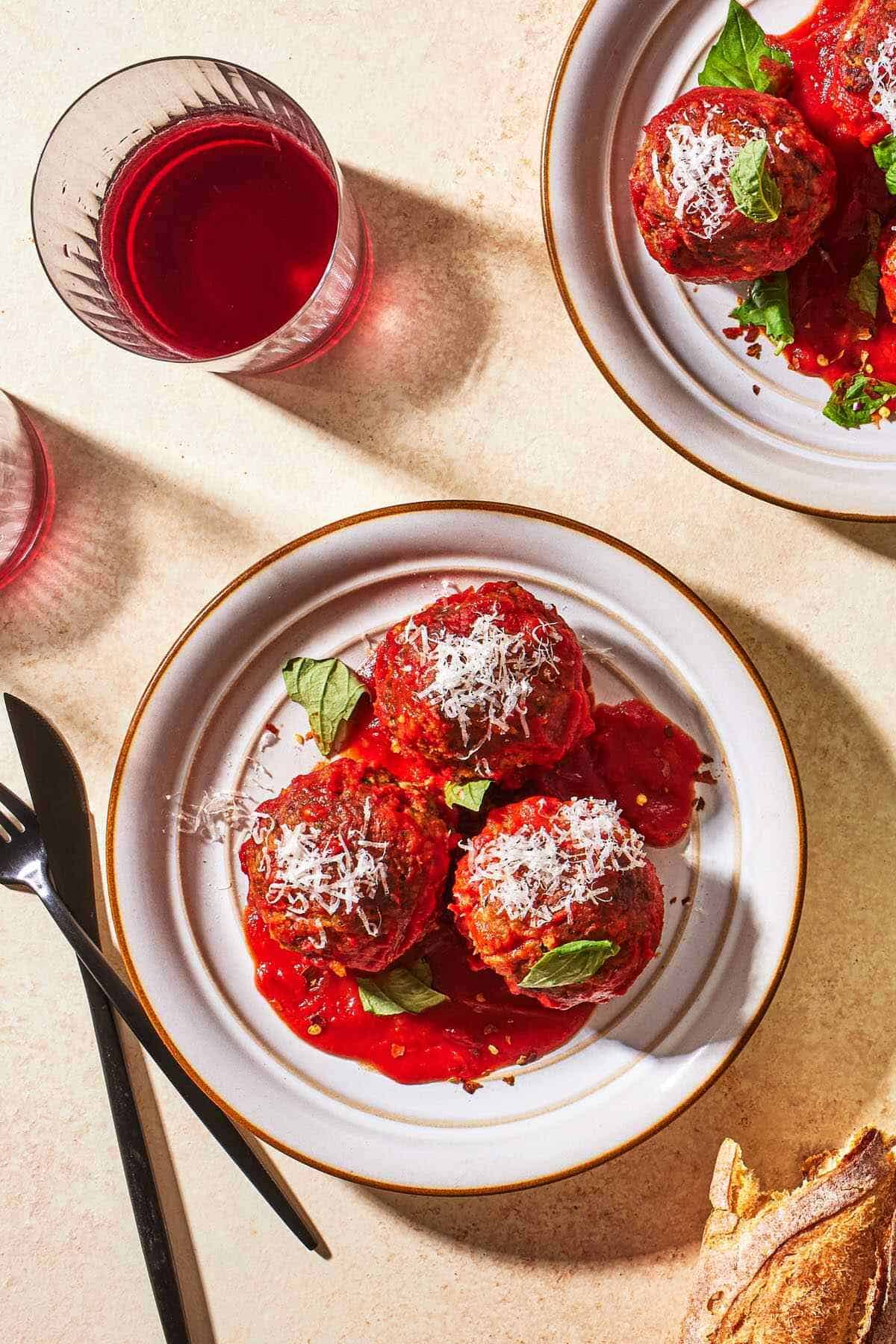 an overhead photo of two plates of three meatballs each with sauce garnished with basil and grated cheese next to a knife, some crusty bread and a drink in a glass.