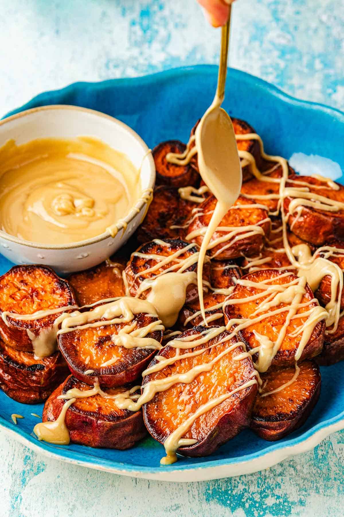 A spoon drizzling honeyed tahini over roasted sweet potatoes, showing its thick and sticky caramel-like consistency.