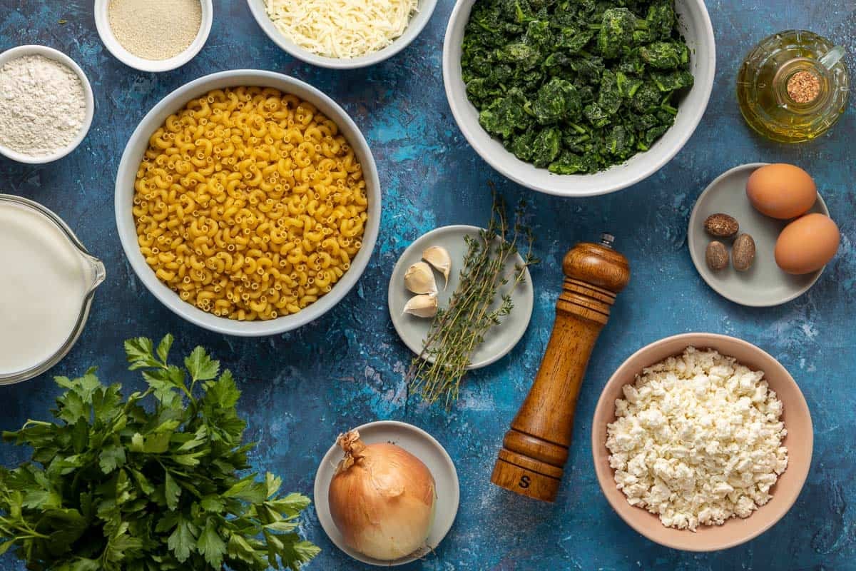 Ingredients for Spanakopita Mac and Cheese including thyme, garlic, onions, eggs, nutmeg, olive oil, feta cheese, parsley, mozzarella, breadcrumbs, and flour.