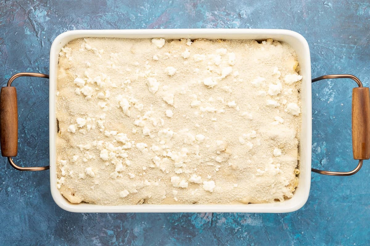 Greek Béchamel Sauce spread on top of pasta in a baking dish with crumbled feta cheese and breadcrumbs on top.