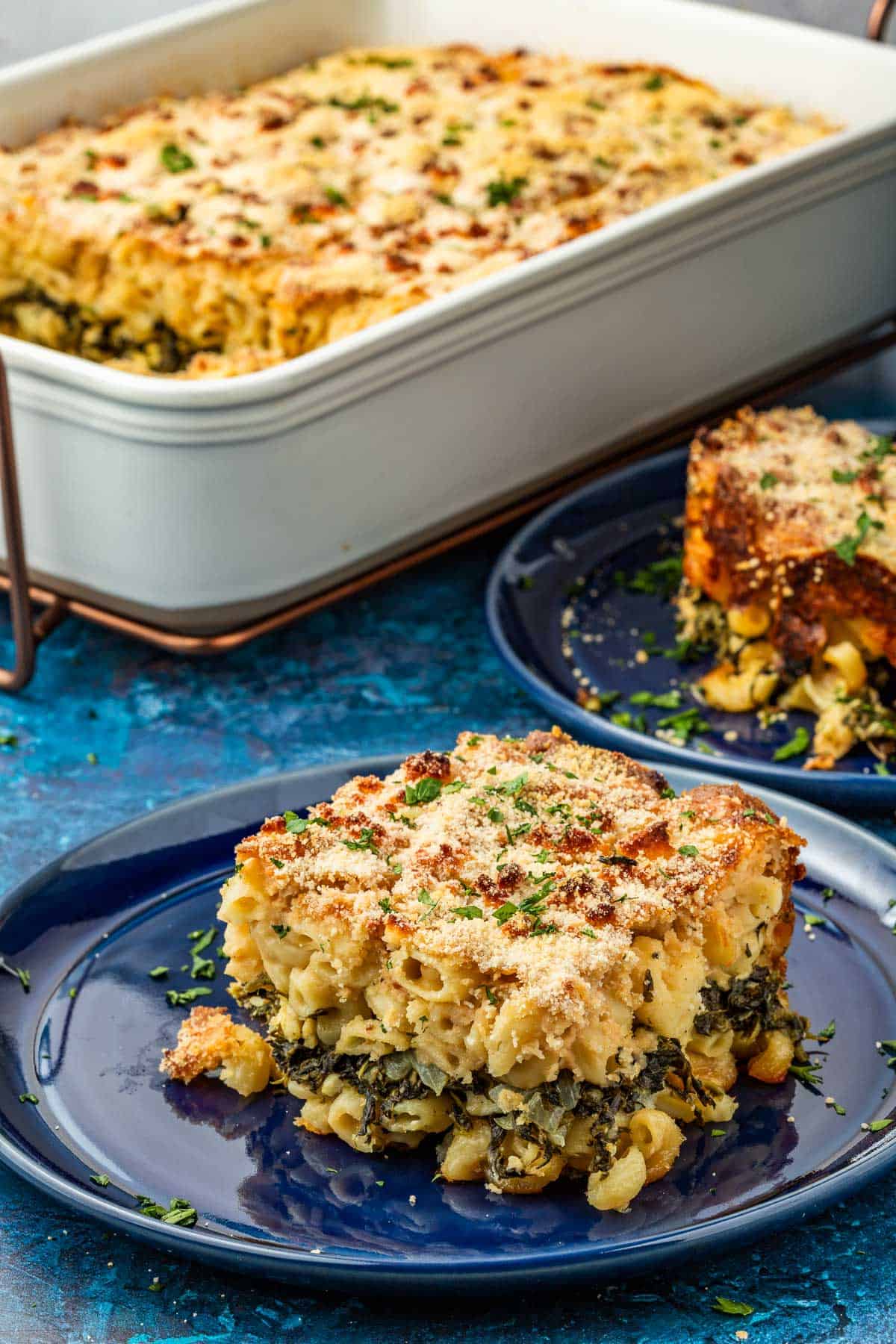 Piece of Spanakopita Mac and cheese on a blue plate, showing the layer of spinach, onion, and herb mixture in the center and the golden brown curst. The casserole dish is in the background.