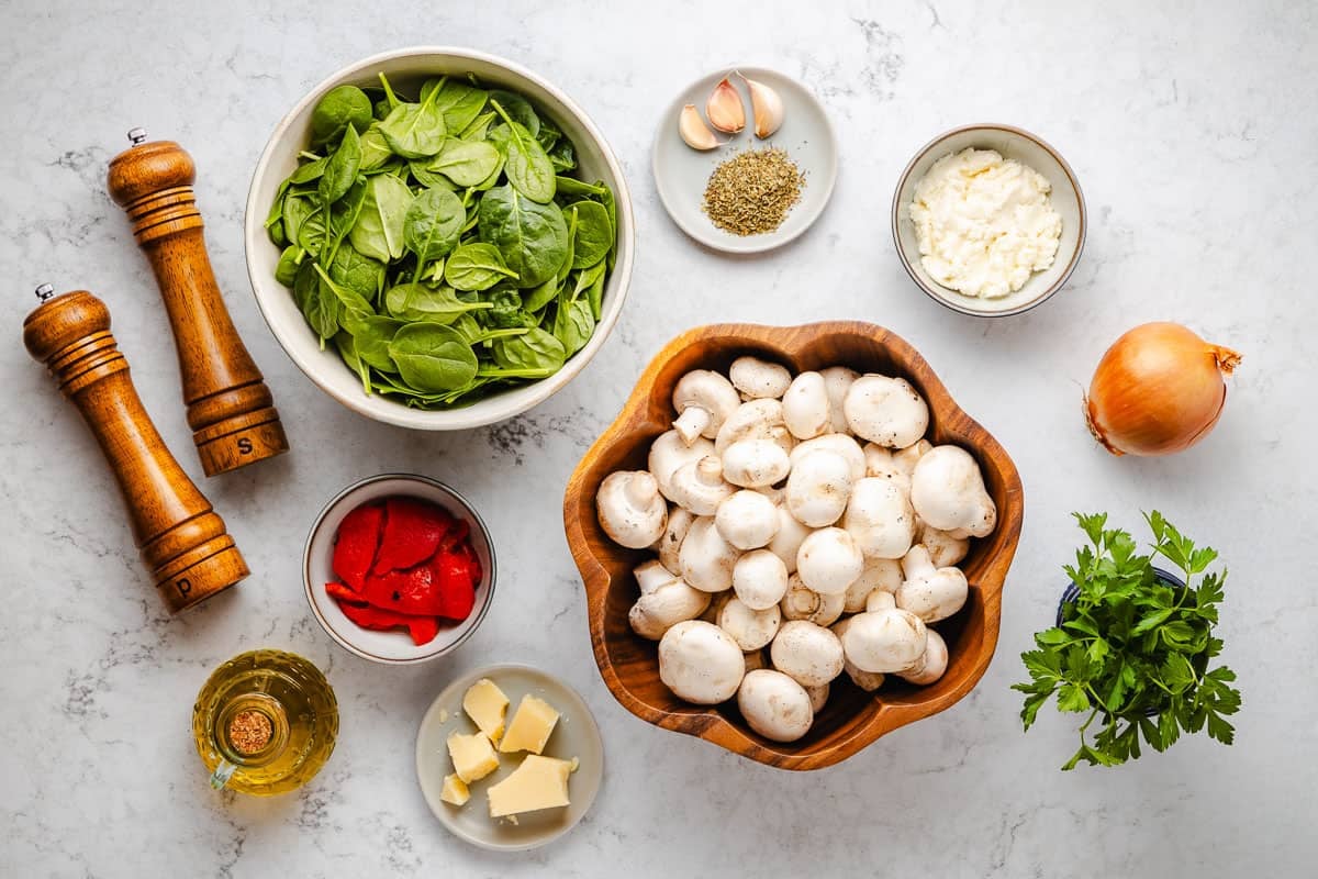 ingredients for stuffed mushrooms including mushrooms, olive oil, salt, pepper, garlic, baby spinach, roasted red peppers, onion, italian seasoning, ricotta cheese, parsley and parmesan cheese.