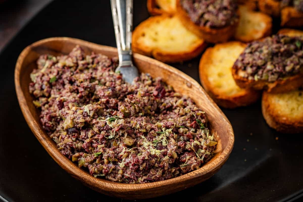 a close up of olive tapenade in a small bowl with a spreading knife on a platter with toasted slices of french bread, two with the tapenade.