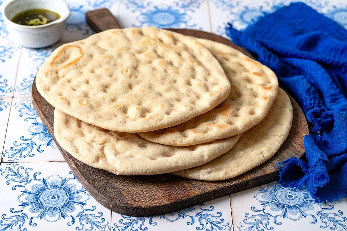 a stack of baked taboon flatbreads on a wooden serving tray next to a small bowl of olive oil and a blue cloth napkin.