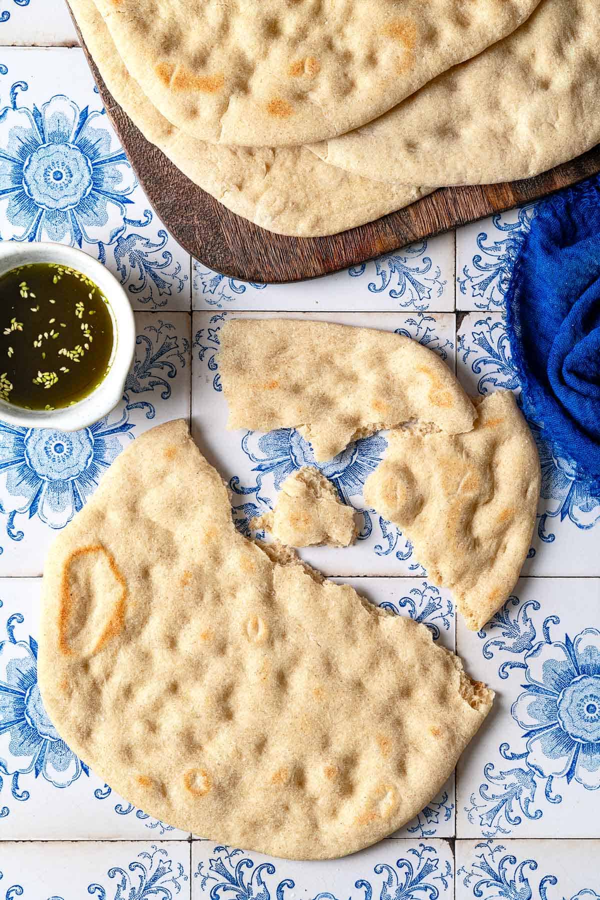 a piece of taboon flatbread torn into large pieces next to seasoned olive oil in a small bowl, and a wooden serving tray with the rest of the taboon.