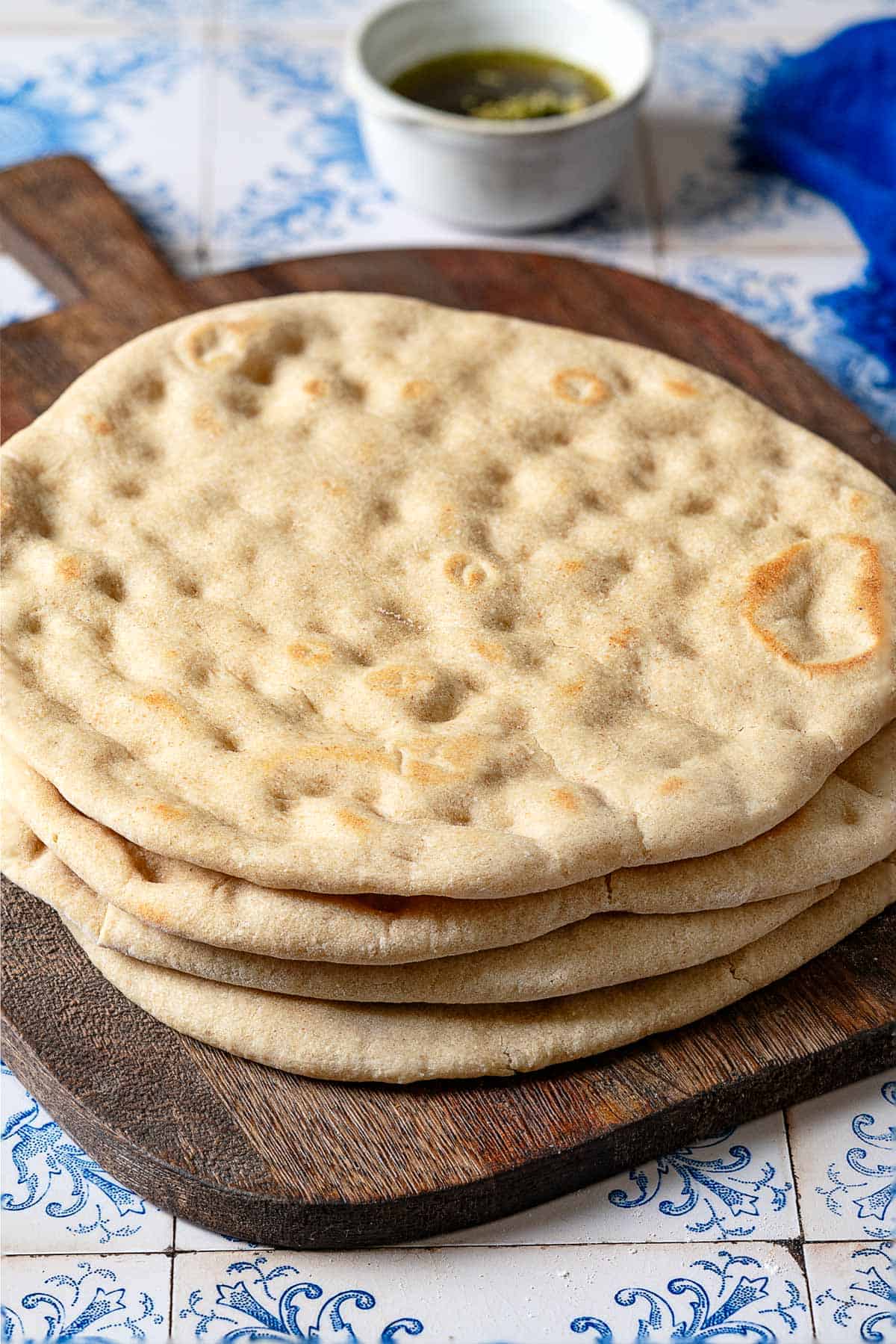 a close up of a stack of baked taboon flatbreads on a wooden serving tray in front of a small bowl of olive oil and a blue cloth napkin.