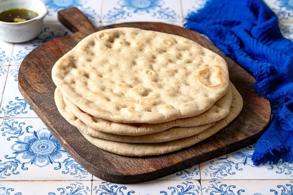 a stack of baked taboon flatbreads on a wooden serving tray next to a small bowl of olive oil and a blue cloth napkin.