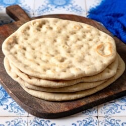 a stack of baked taboon flatbreads on a wooden serving tray next to a small bowl of olive oil and a blue cloth napkin.