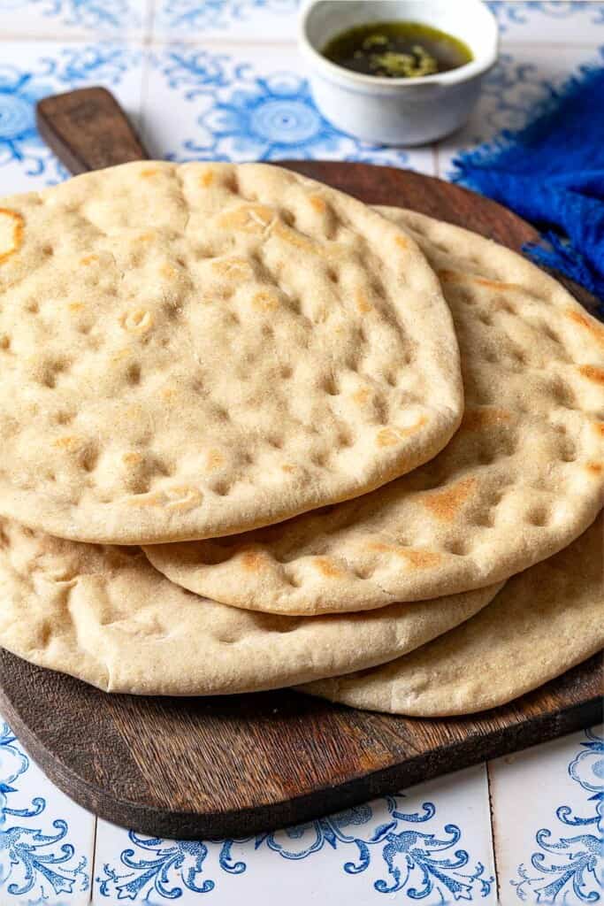 a stack of baked taboon flatbreads on a wooden serving tray in front of a small bowl of olive oil and a blue cloth napkin.