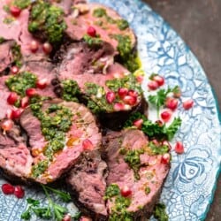 a close up of slices of beef tenderloin topped with chermoula sauce and pomegranate seeds on a serving platter.