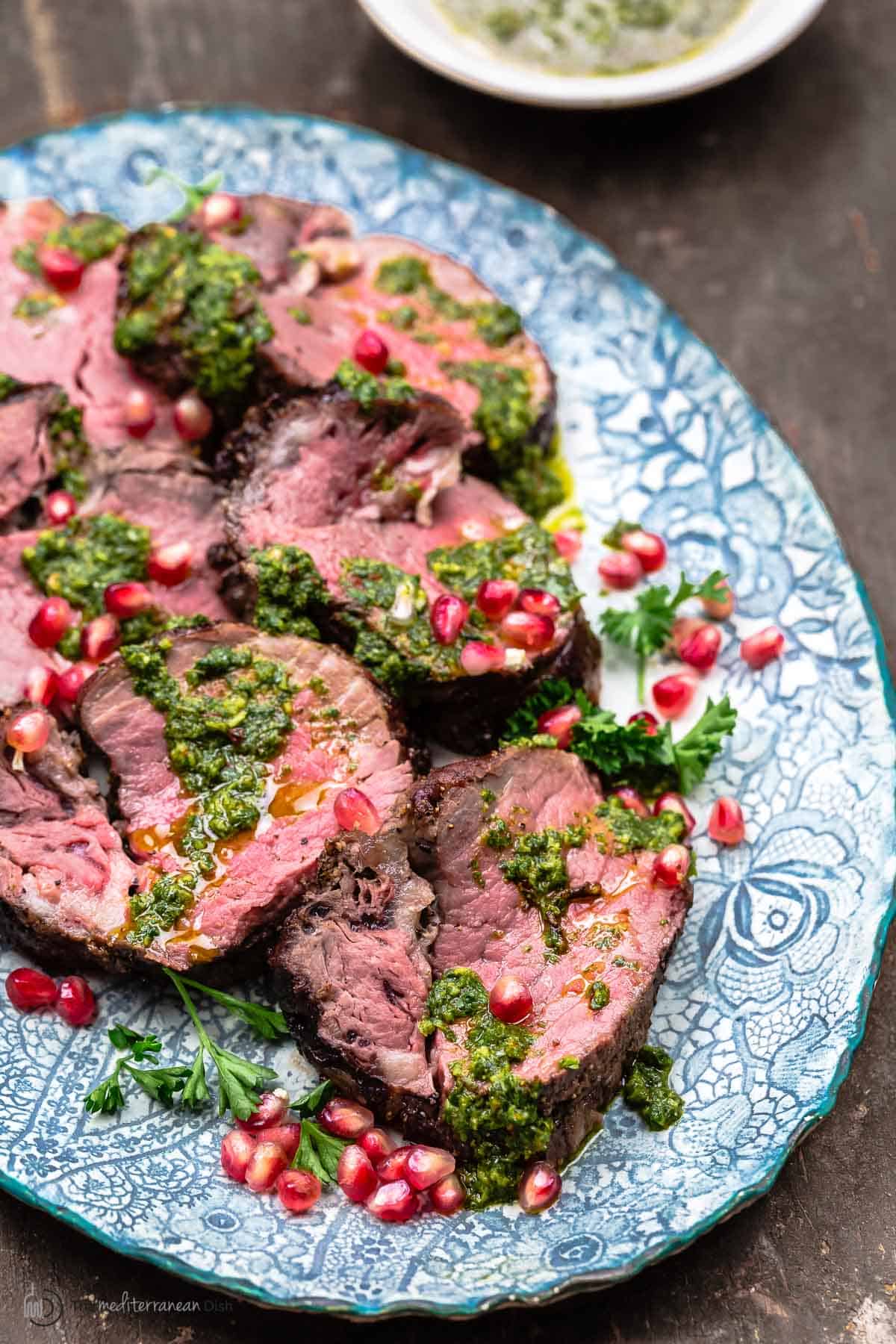 a close up of slices of beef tenderloin topped with chermoula sauce and pomegranate seeds on a serving platter.