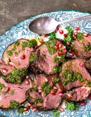 slices of beef tenderloin topped with chermoula sauce and pomegranate seeds on a serving platter with a spoon next to a small bowl of chermoula sauce.