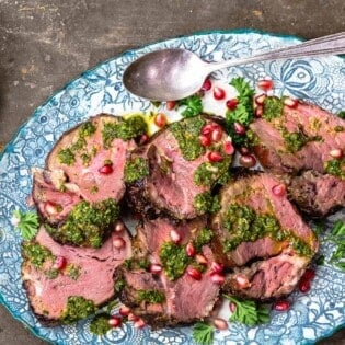 slices of beef tenderloin topped with chermoula sauce and pomegranate seeds on a serving platter with a spoon next to a small bowl of chermoula sauce.