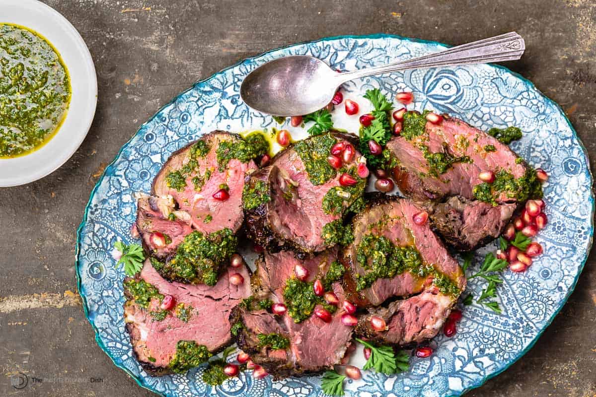 slices of beef tenderloin topped with chermoula sauce and pomegranate seeds on a serving platter with a spoon next to a small bowl of chermoula sauce.