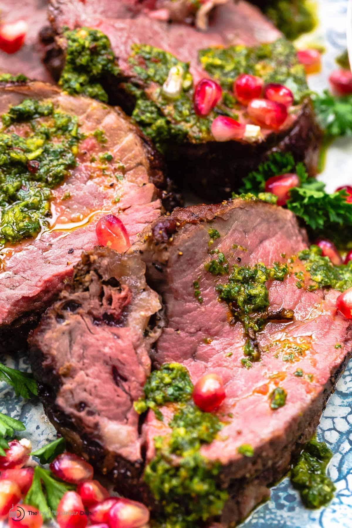 a close up of slices of beef tenderloin topped with chermoula sauce and pomegranate seeds.