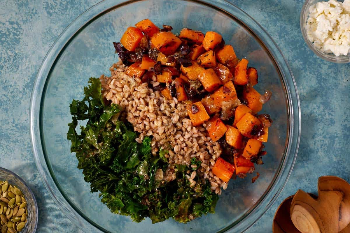 the roasted vegetables, kale and farro in a bowl, prior to mixing together next to small bowls of crumbled goat cheese and pepitas.