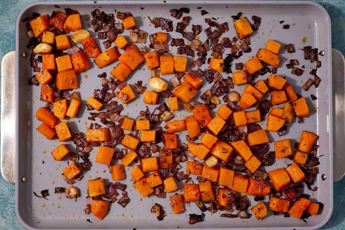 cubed butternut squash, chopped red onion and cloves of garlic on a baking sheet after roasting.