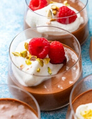 a close up of 2 glass cups of olive oil chocolate mousse topped with raspberries, whipped cream and nuts.