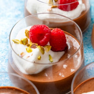 a close up of 2 glass cups of olive oil chocolate mousse topped with raspberries, whipped cream and nuts.