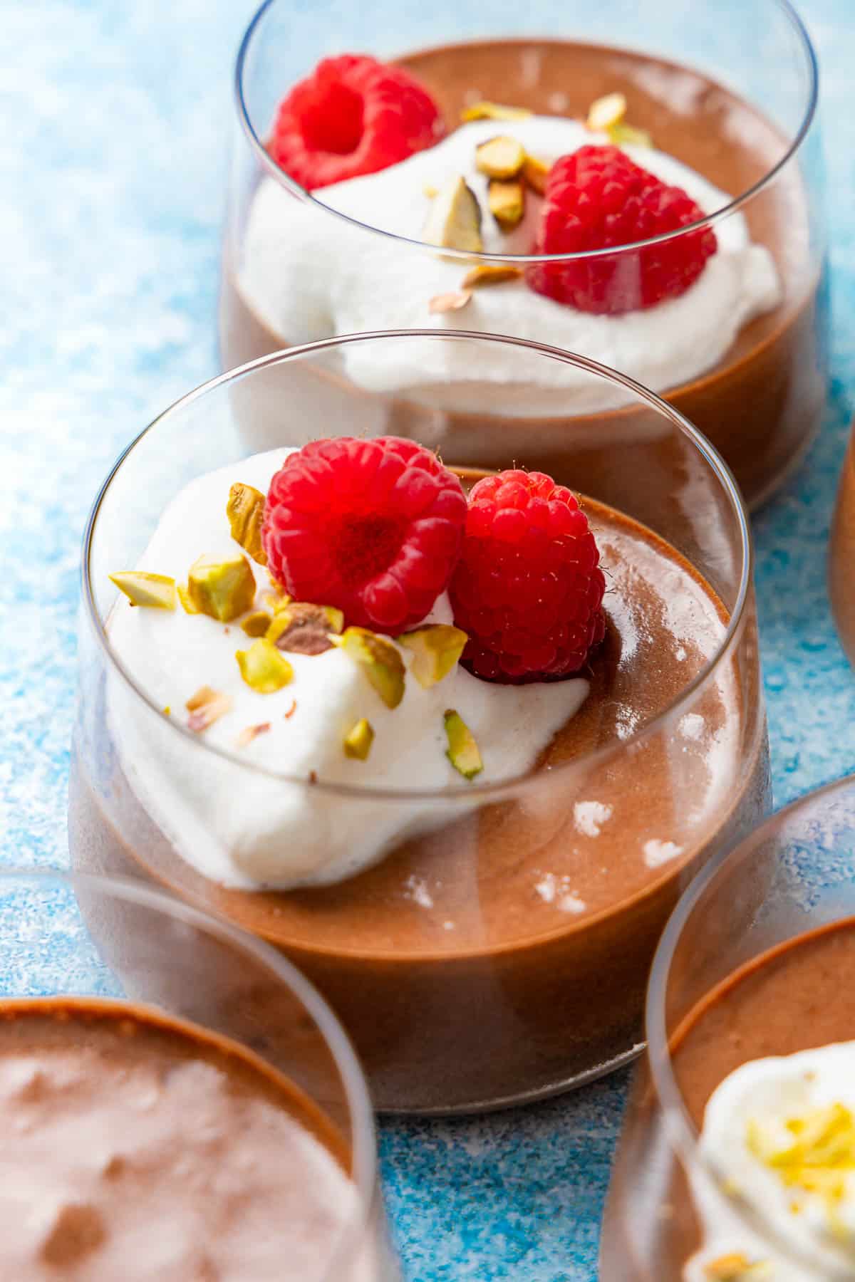 a close up of 2 glass cups of olive oil chocolate mousse topped with raspberries, whipped cream and nuts.