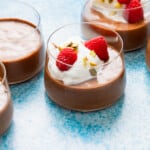 a close up of 3 glass cups of olive oil chocolate mousse, some topped with raspberries, whipped cream and nuts.