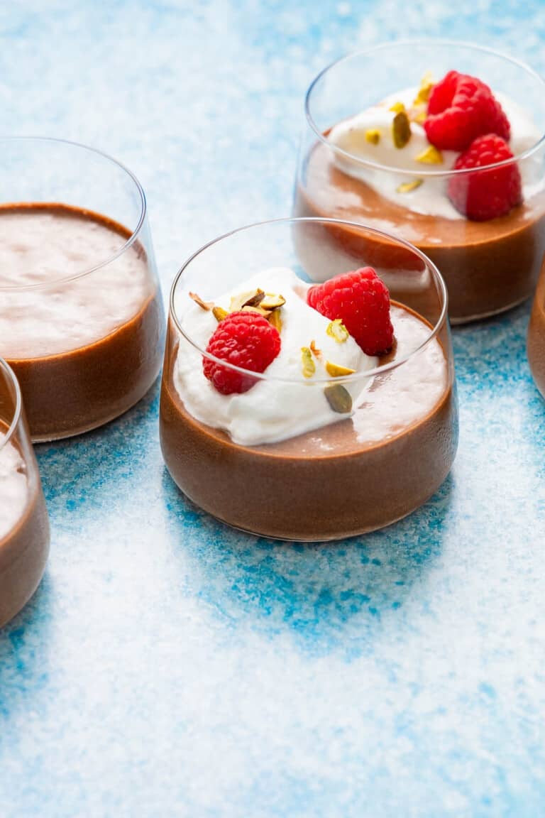 a close up of 3 glass cups of olive oil chocolate mousse, some topped with raspberries, whipped cream and nuts.