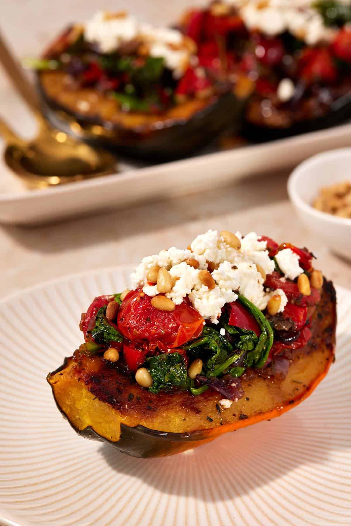 One stuffed acorn squash quarter on a plate, showing the tender squash, burst tomatoes, wilted spinach, golden brown pine nuts, and crumbled goat cheese.
