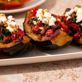 Side shot of three roasted acorn squash quarters stuffed with spinach, onions, and tomatoes and topped with goat cheese and pine nuts.