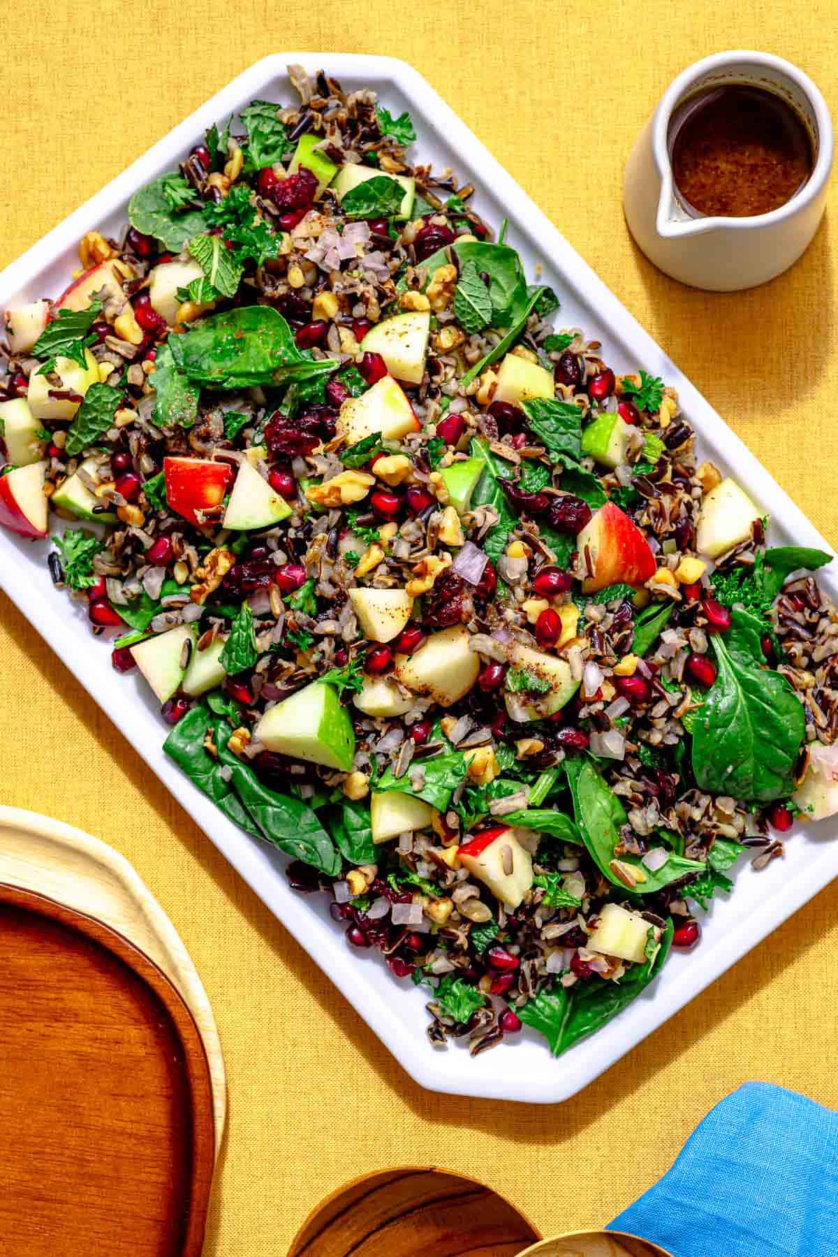 overhead shot of wild rice salad on a rectangular serving platter with a small spouted pitcher of dressing on the side.